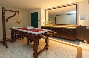 Uma cama de madeira tradicional com colcha vermelha e branca, uma mesa de cabeceira de madeira, um espelho grande e uma porta verde num quarto de hotel.
