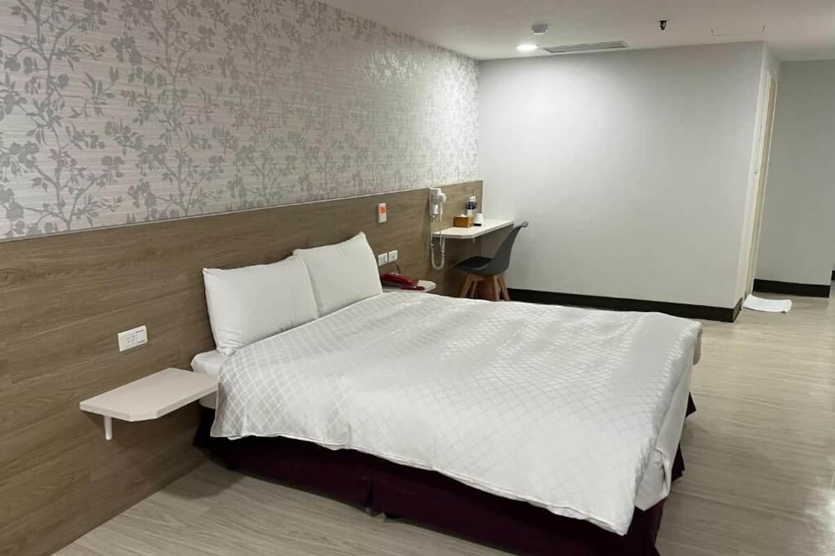 A hotel room with a large bed, a small desk, a chair, and a shower area.