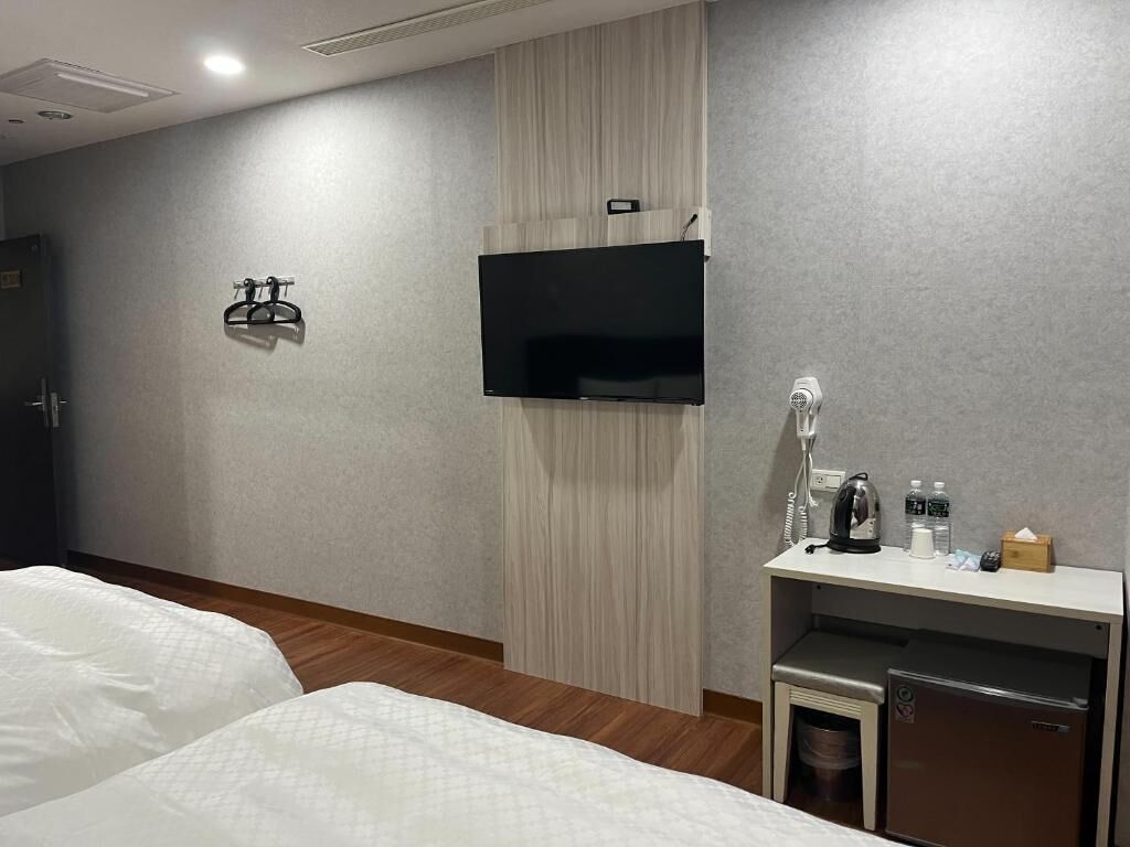 A hotel room with two beds, a flat-screen TV mounted on the wall, a small desk with a kettle, and a mini-fridge.