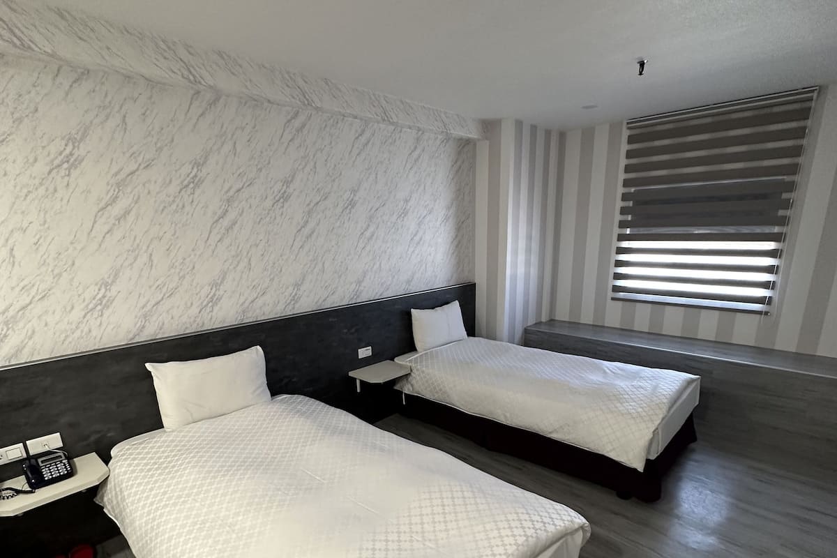 A modern hotel room with two beds, a marble wall, a wooden floor, a nightstand with a phone, and a window with blinds.