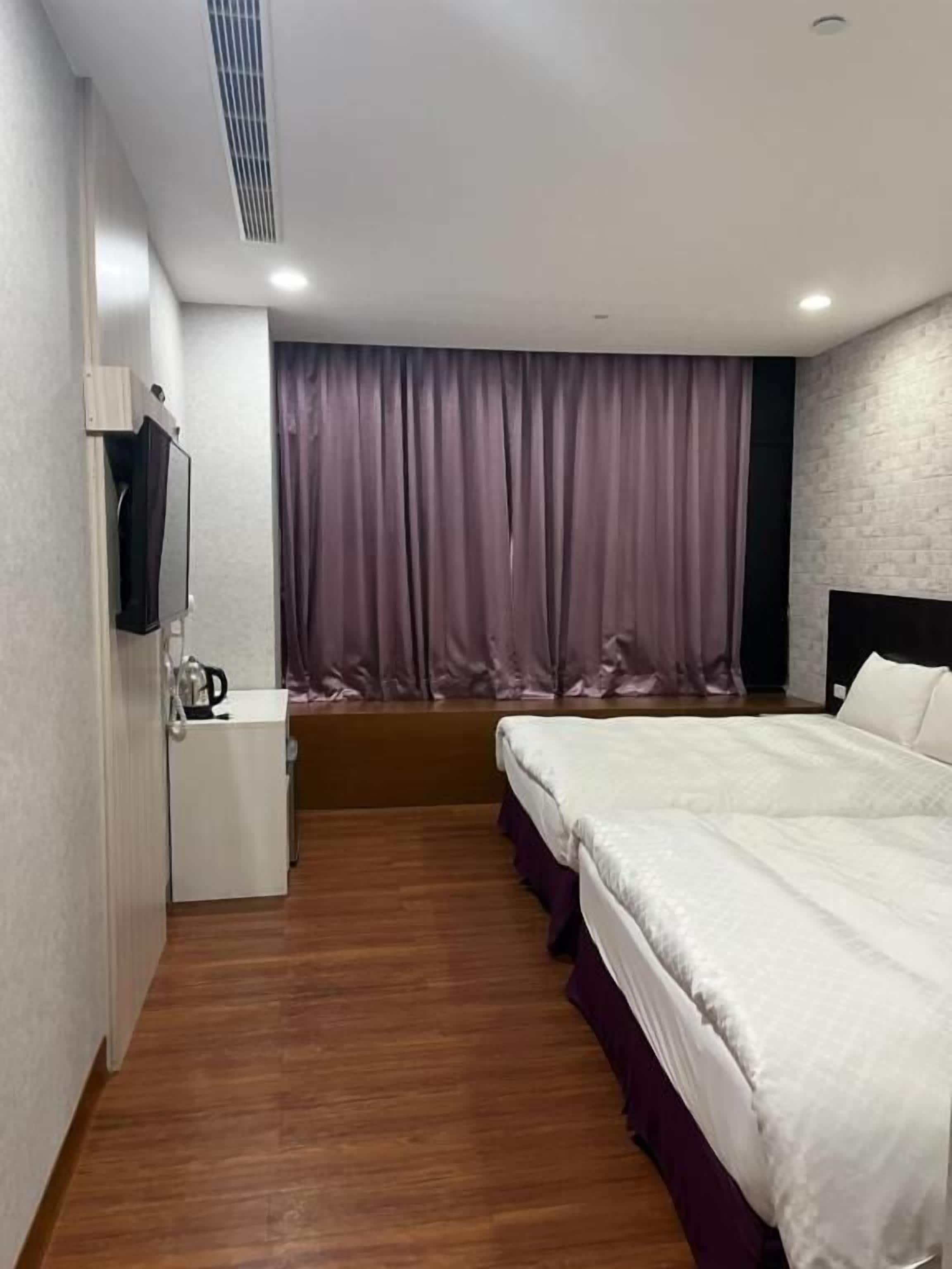 Habitación de hotel con cama, mesita de noche pequeña, televisor montado en la pared y ventana con cortinas.