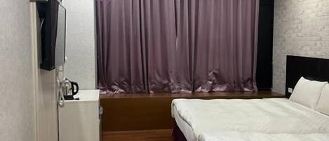 Habitación de hotel con cama, mesita de noche pequeña, televisor montado en la pared y ventana con cortinas.
