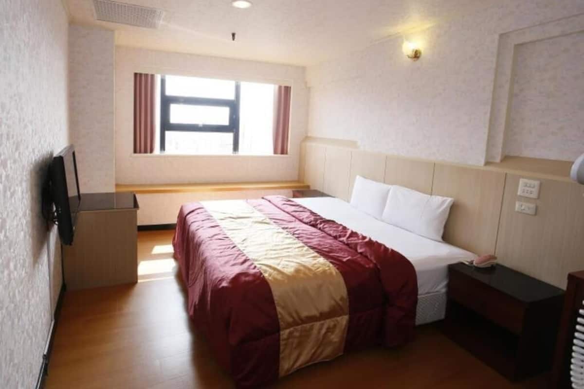 A hotel room with a bed, a desk, a chair, a window with curtains, and a wall-mounted TV.