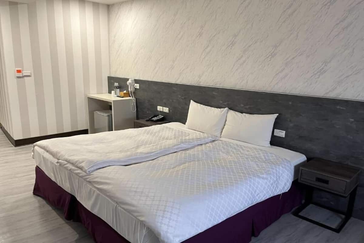 A modern hotel room with a large bed, a bedside table, a small desk, and a chair.