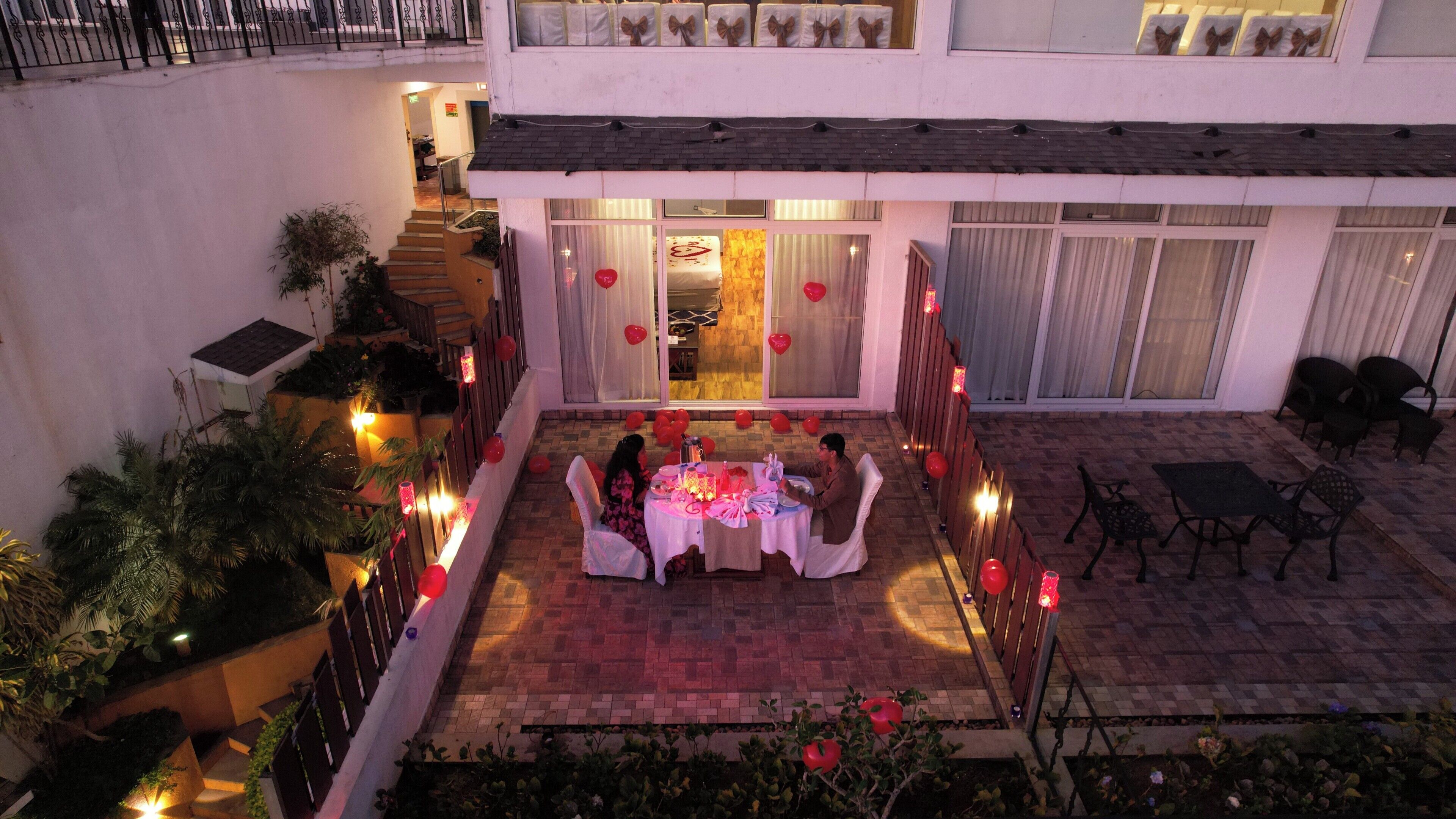 Un espace extérieur aménagé pour prendre les repas, avec une table dressée pour deux personnes, entourée de plantes en pot et de ballons rouges.