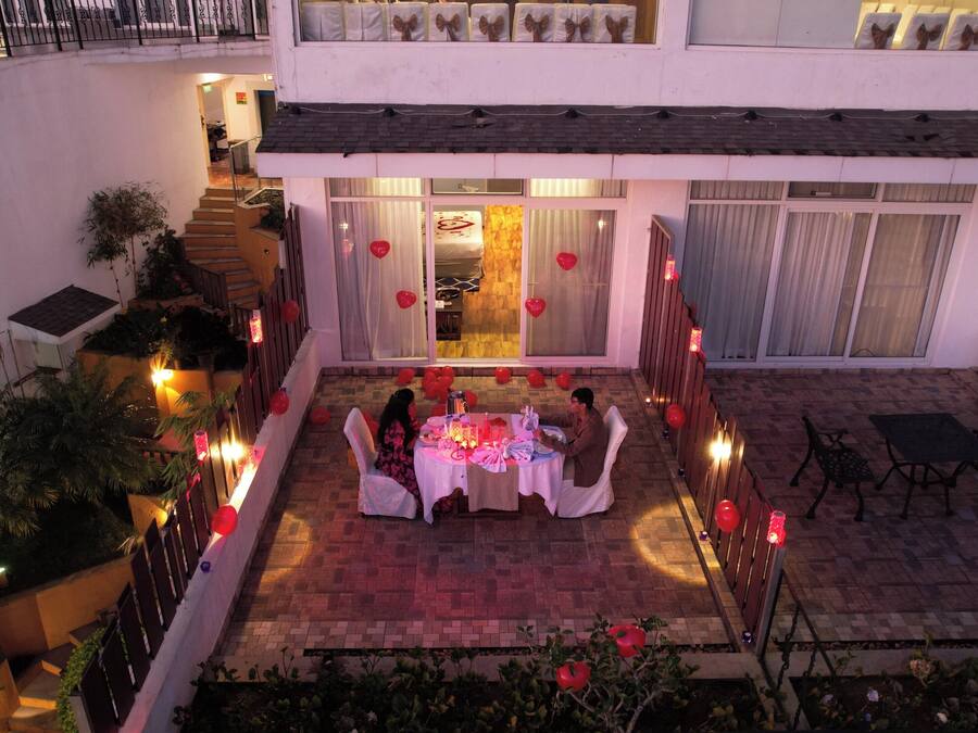 Un espace extérieur aménagé pour prendre les repas, avec une table dressée pour deux personnes, entourée de plantes en pot et de ballons rouges.
