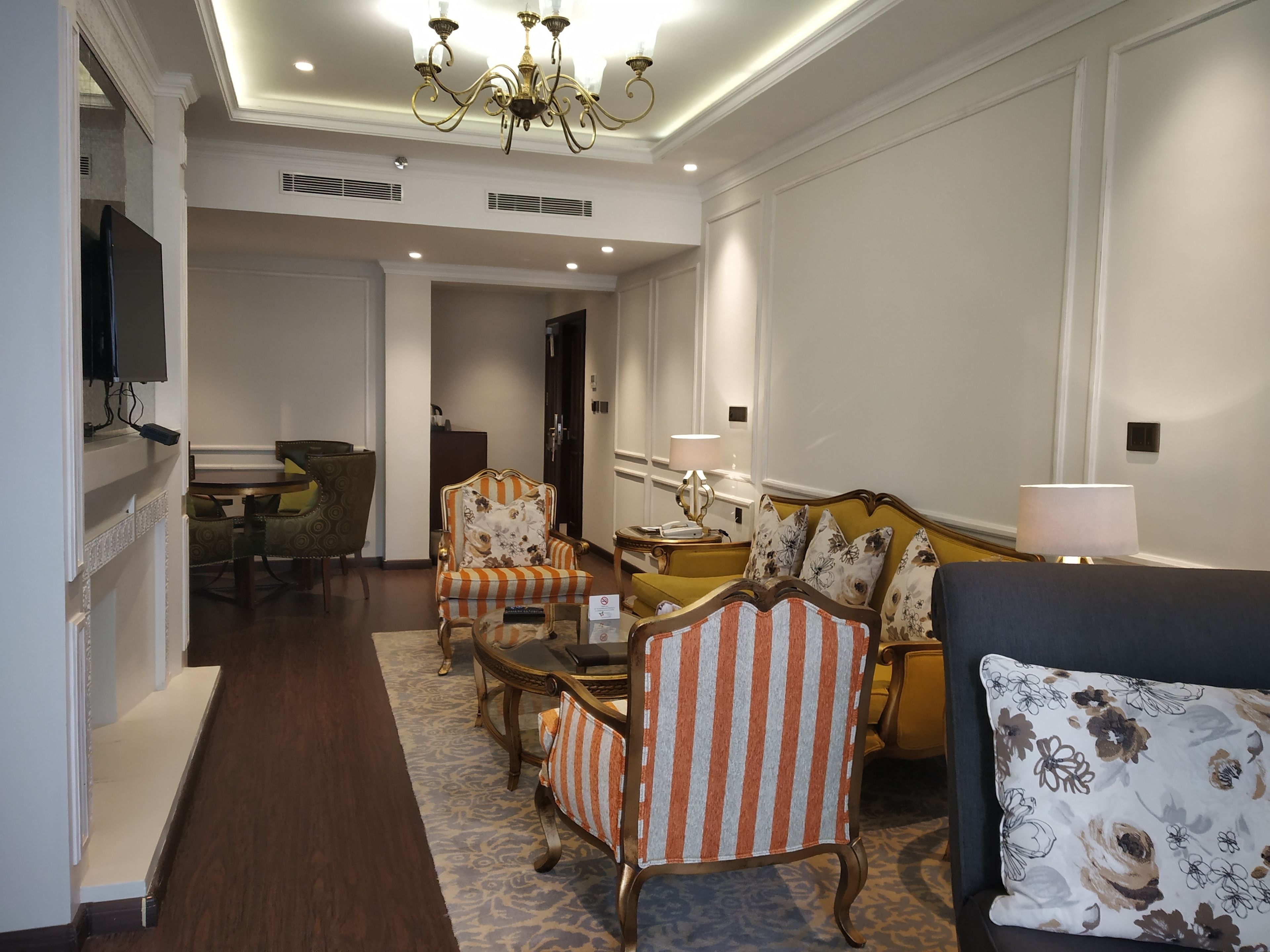 Un salon spacieux agrémenté d’un lustre, d’un canapé, de fauteuils et d’un téléviseur fixé au mur.