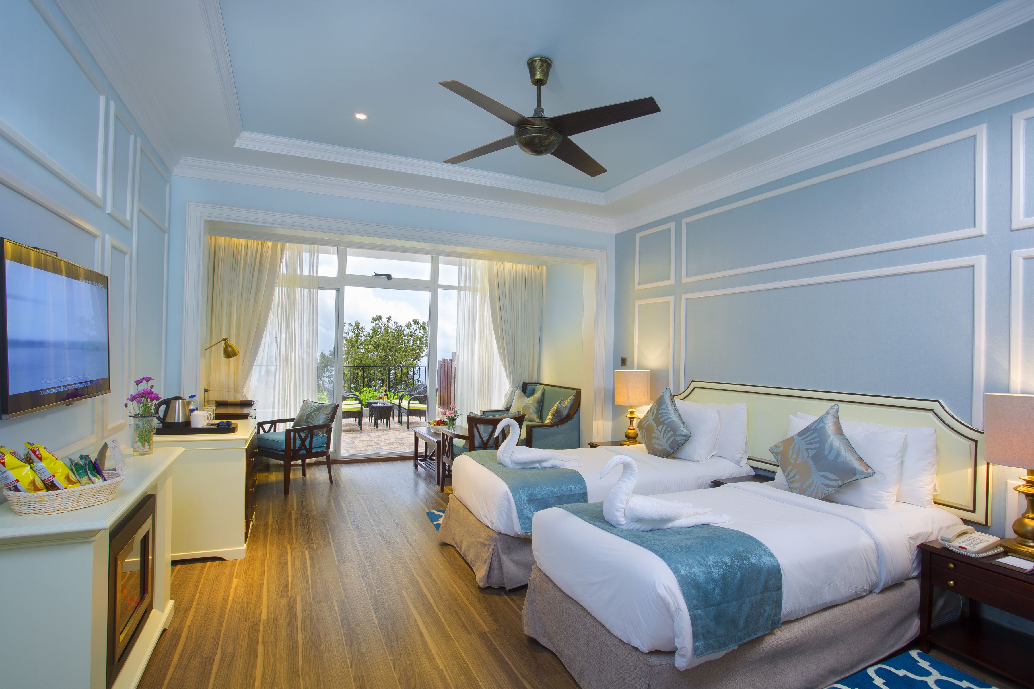 A hotel room with two beds, a TV, a desk with a chair, a ceiling fan, and a view of the outdoors.
