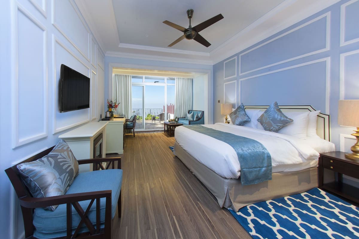 A hotel room with a bed, a desk, a chair, a television, and a balcony with curtains.