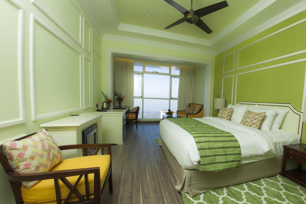 A hotel room with a large bed, a desk, a chair, and a view of the city through large windows.
