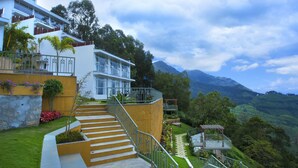 Un hotel moderno con una escalera que lleva a un balcón, rodeado de abundante vegetación y montañas al fondo.