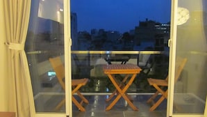 Un balcon avec une table et deux chaises, un rideau et une vue sur les bâtiments au crépuscule.