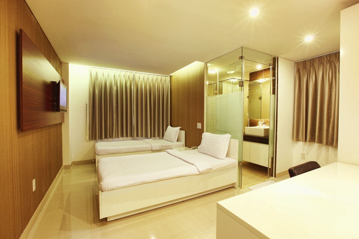 Une chambre d’hôtel avec deux lits, un bureau, un miroir et une salle de bain visible à travers une paroi en verre.