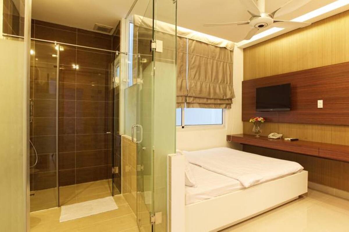 Una habitación de hotel moderna con ducha de vidrio, una cama, una mesita de noche con televisor y un panel de pared de madera.