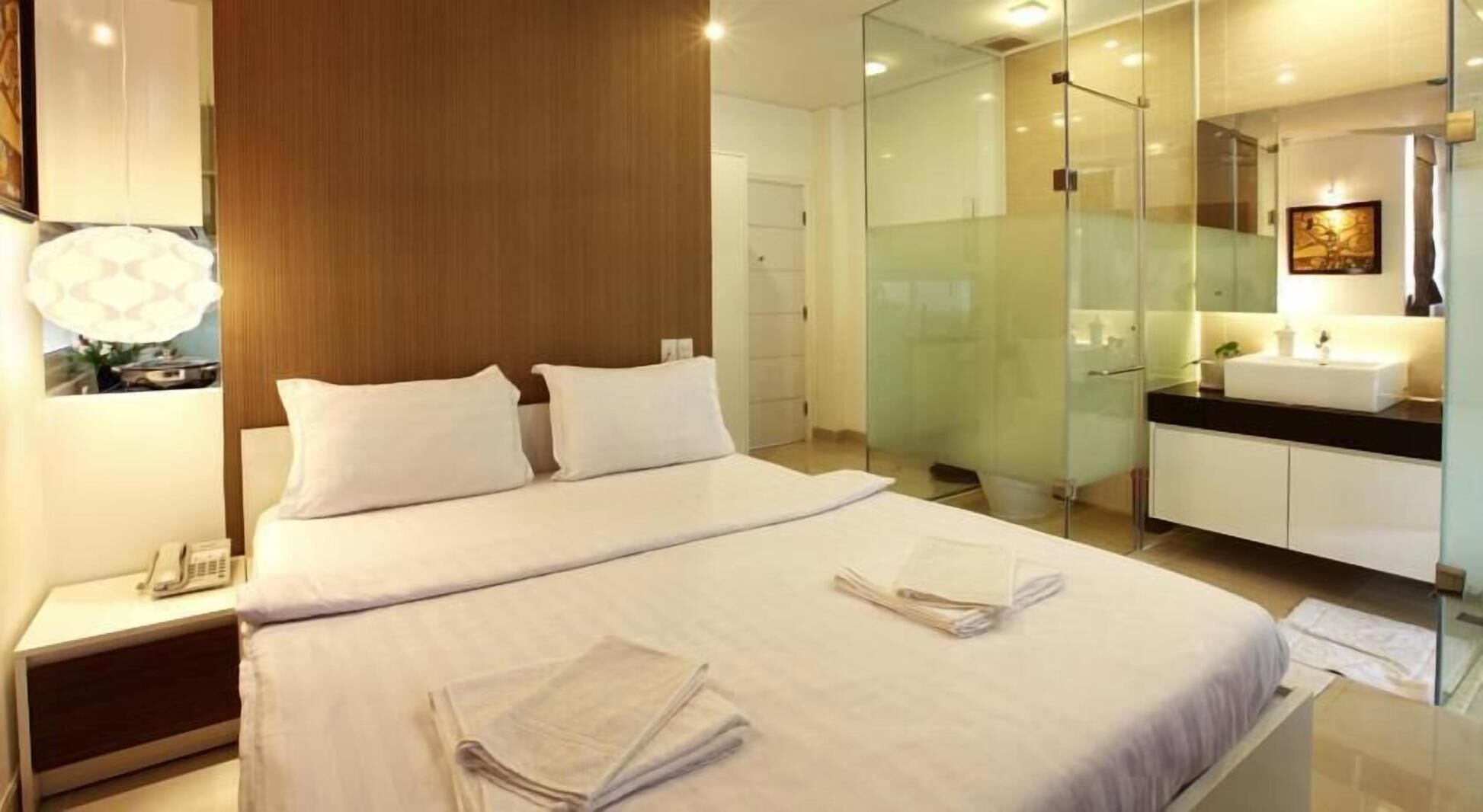 Una habitación de hotel moderna con una cama grande, una mesita de noche con teléfono, ducha con mampara de vidrio y un baño con lavamanos y espejo.