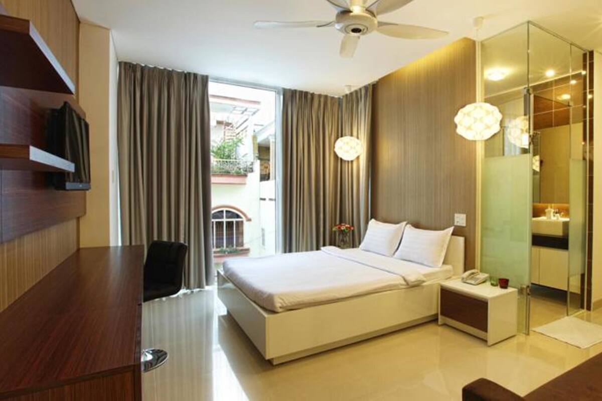 Una habitación de hotel moderna con una cama grande, un escritorio, una silla, un ventilador de techo y un baño con mampara de vidrio.