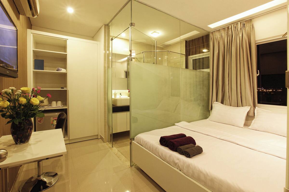 Habitación de hotel con cama, escritorio, silla, cabina de ducha de vidrio y vista nocturna de la ciudad.