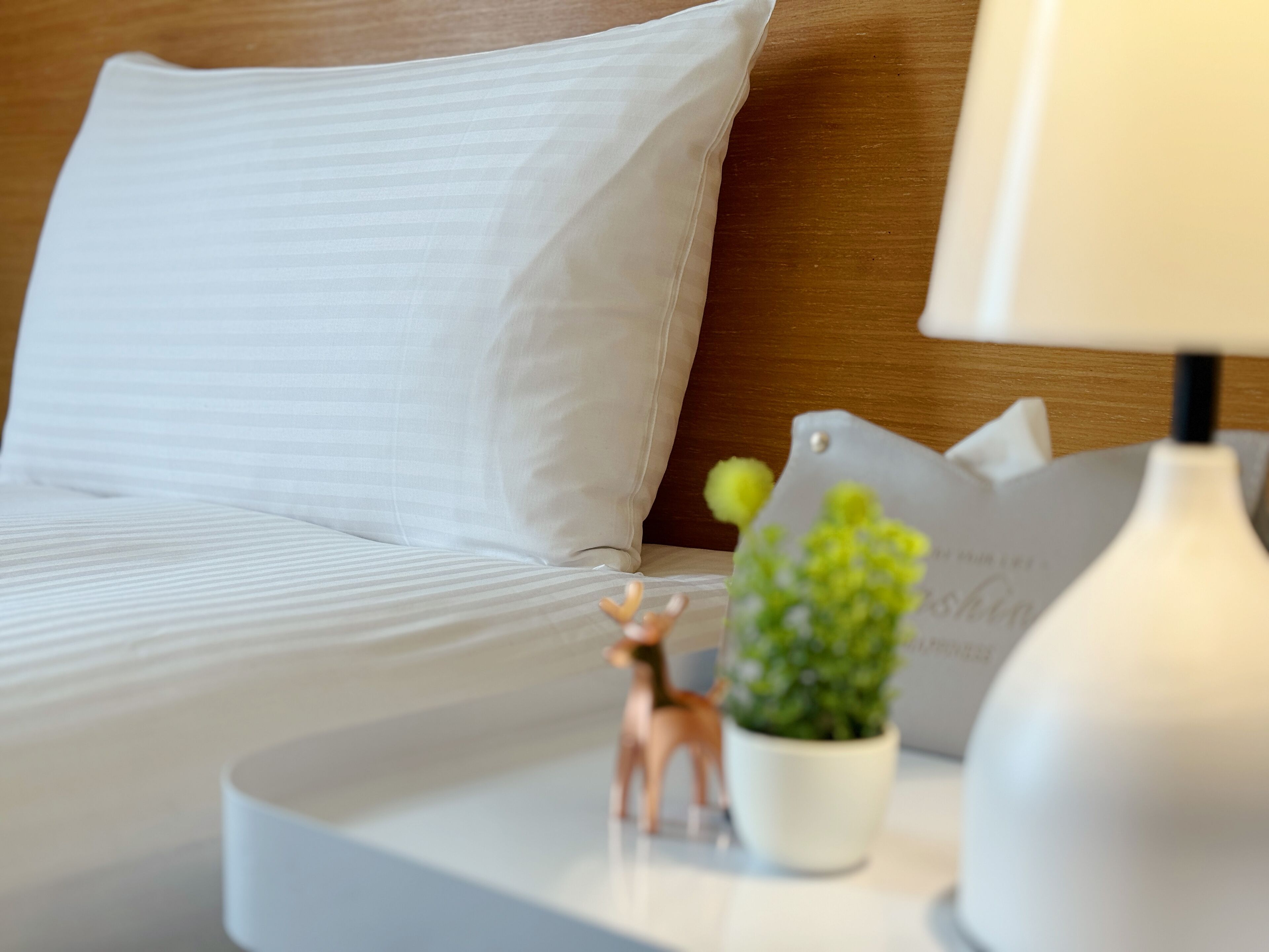 A neatly made bed with white linens, a small potted plant, a decorative deer figurine, and a white lamp on a bedside table.