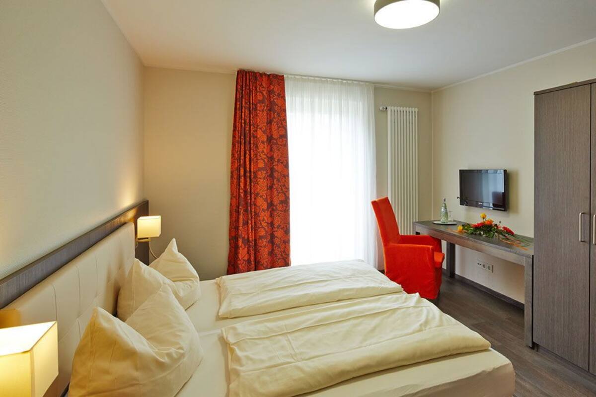 A hotel room with a bed, bedside lamps, a desk with a chair, a television, and a wardrobe.