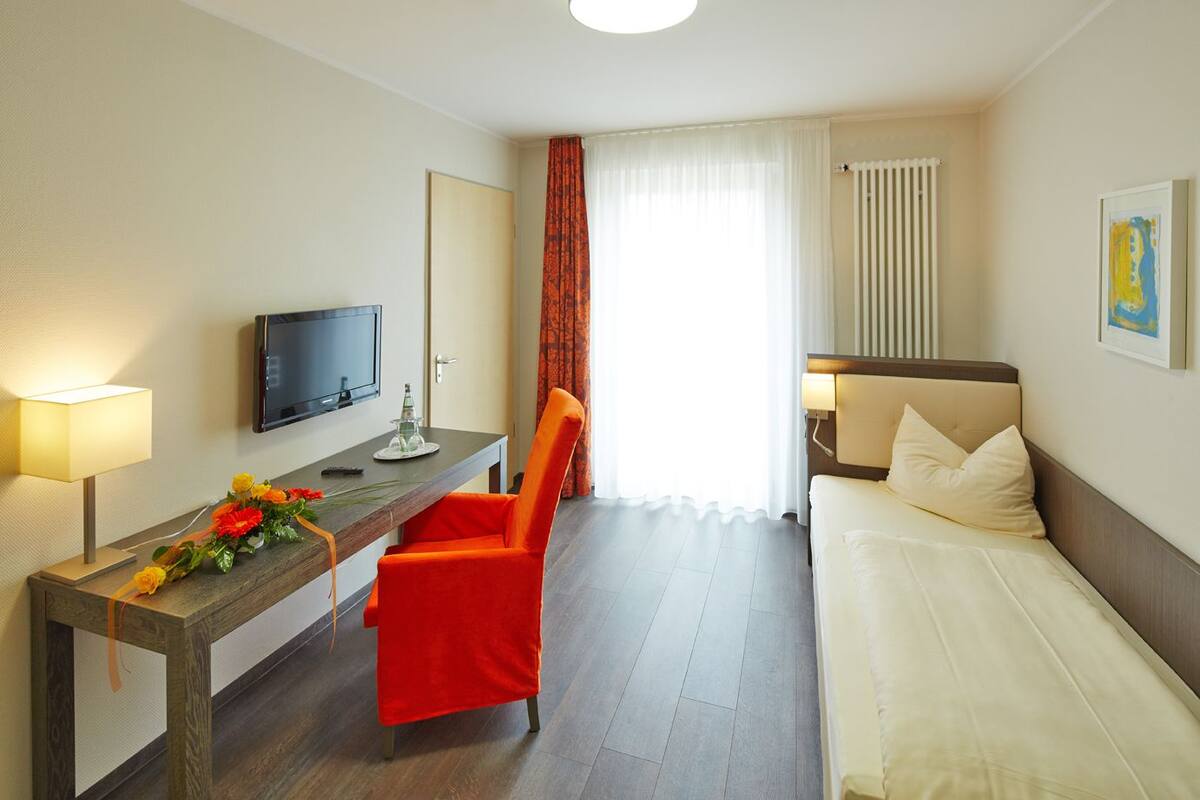 A hotel room with a bed, a desk with a chair, a television, and a window with curtains.