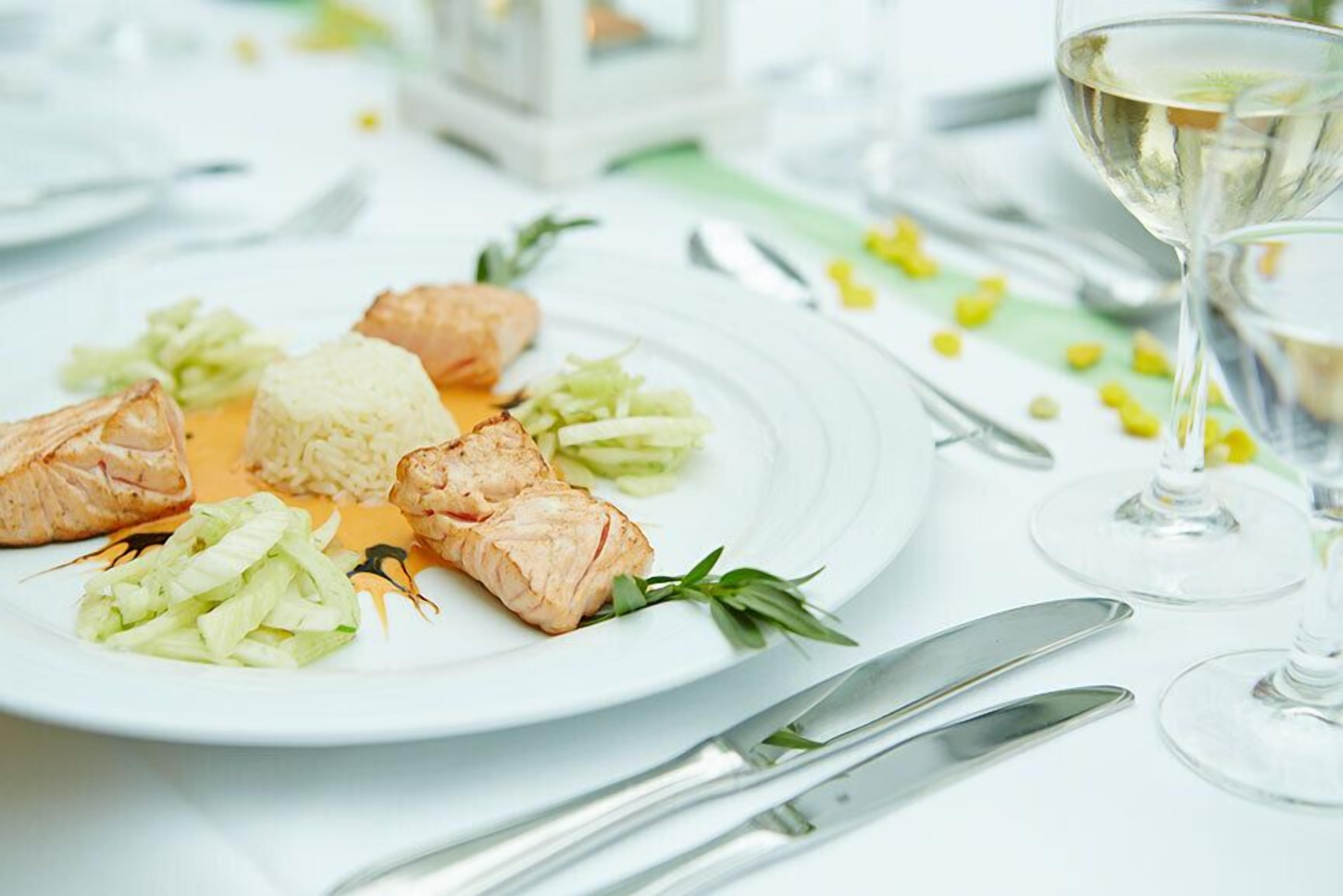 Ein Teller mit Lachs, Reis und Gemüse auf einem weißen Tisch, mit Besteck und einem Glas Weißwein.