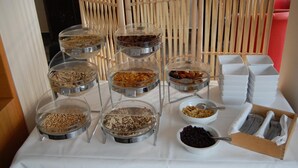 A buffet setup with various food items in clear containers, including cereals, nuts, and fruits, with serving utensils and plates arranged on a table. - Hotel FIT (Much)