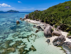 A tropical beach with clear turquoise waters, large boulders, and dense palm trees.