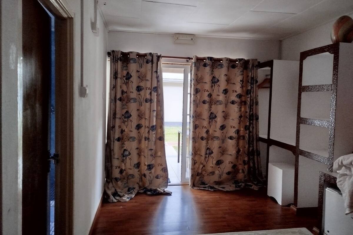Um corredor com piso de madeira, uma porta com faixa azul e vista para o exterior através de uma porta com cortinas estampadas.
