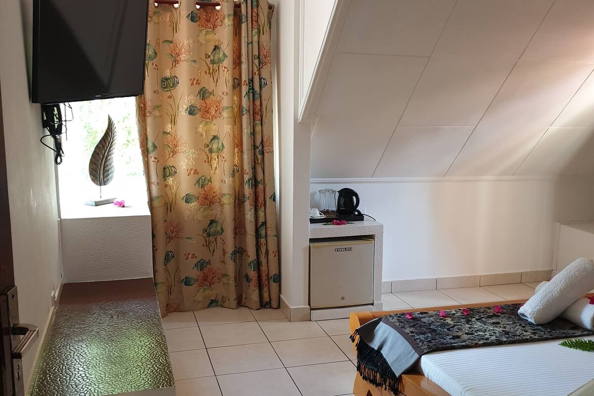 Quarto pequeno e bem iluminado, com uma cama, uma escrivaninha e uma janela com cortinas.