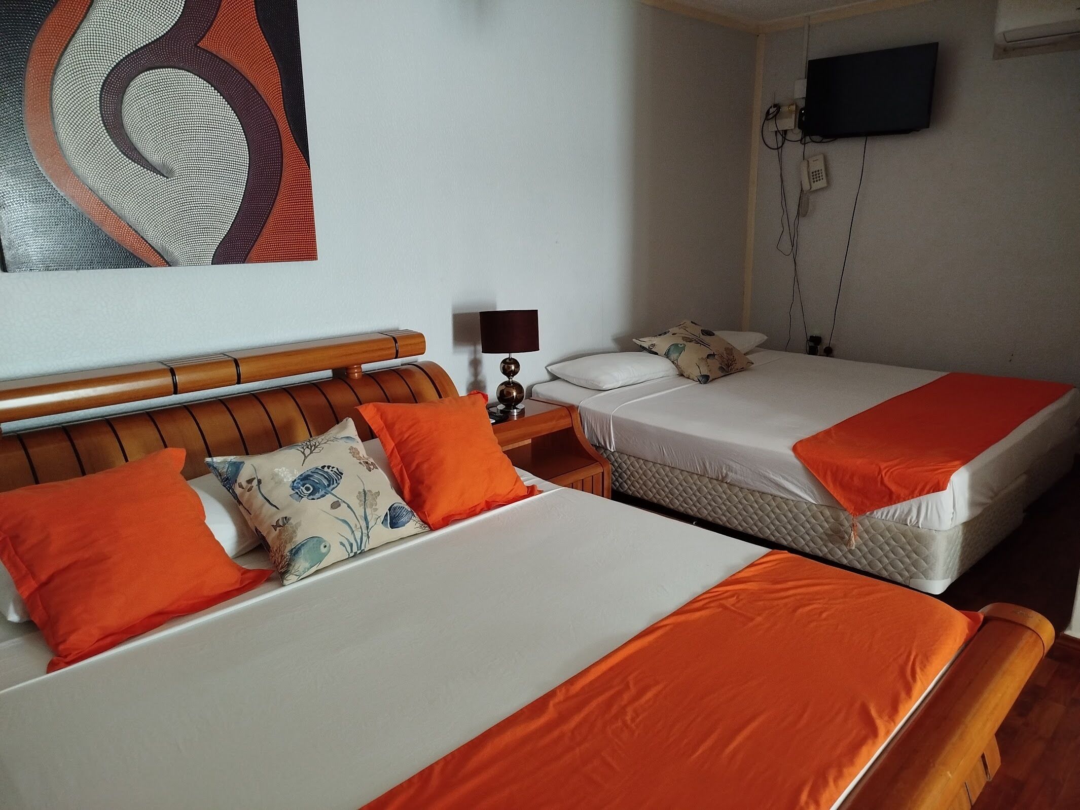 A hotel room with two beds, each with orange bedding and decorative pillows. There is a wooden headboard, a bedside table with a lamp, and a wall-mounted TV.