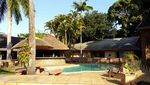 A pool area with a thatched roof structure, palm trees, and a brick building. - iLanga Lodge (St. Lucia)