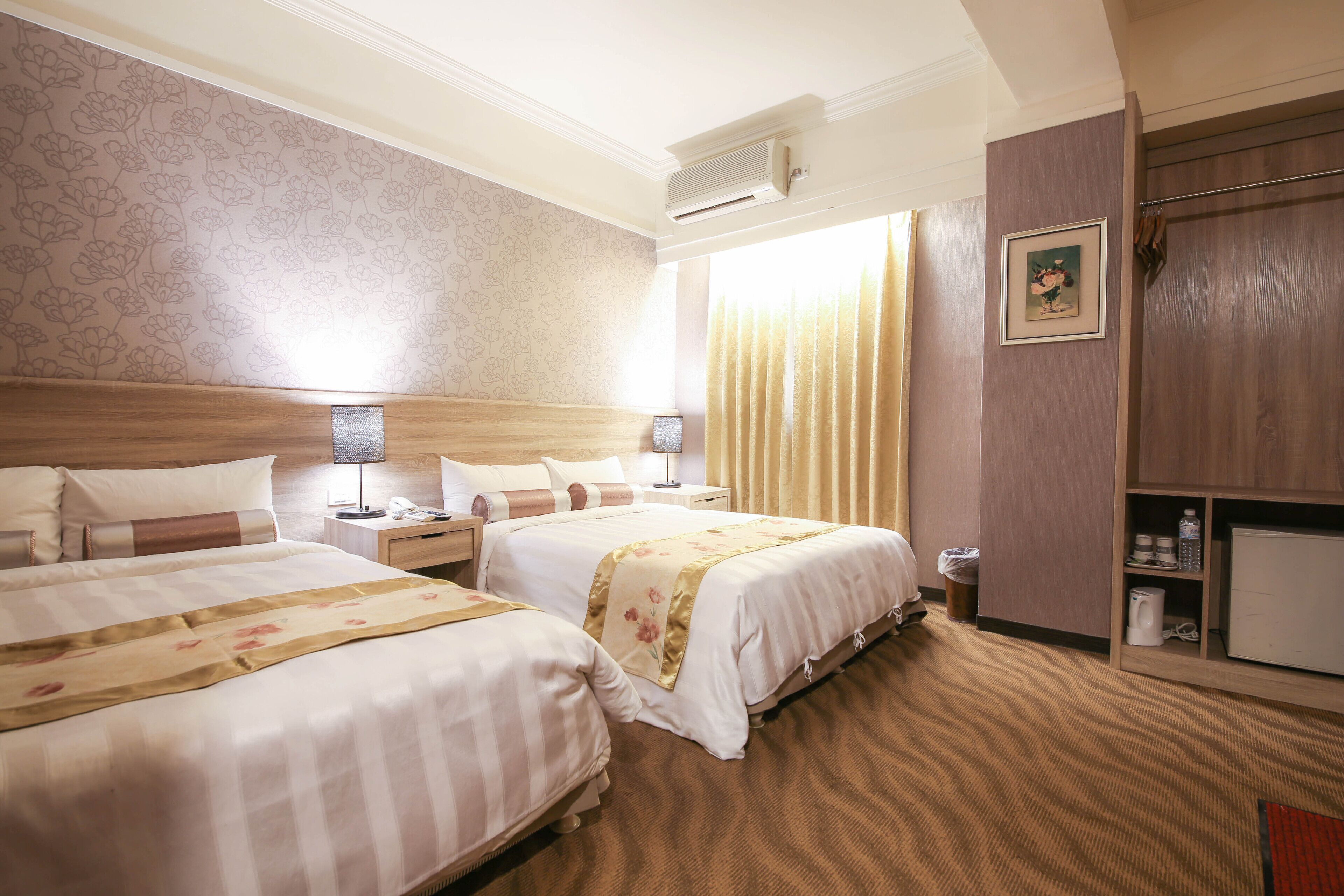 Ein Hotelzimmer mit zwei Betten, einem hölzernen Kopfteil, einem Nachttisch mit Lampe, einem kleinen Tisch mit Telefon, einem Mülleimer, einem Schrank mit Mikrowelle und einem Fenster mit Vorhängen.
