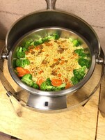 A stainless steel pot with a lid, containing noodles and vegetables, placed on a marble countertop.