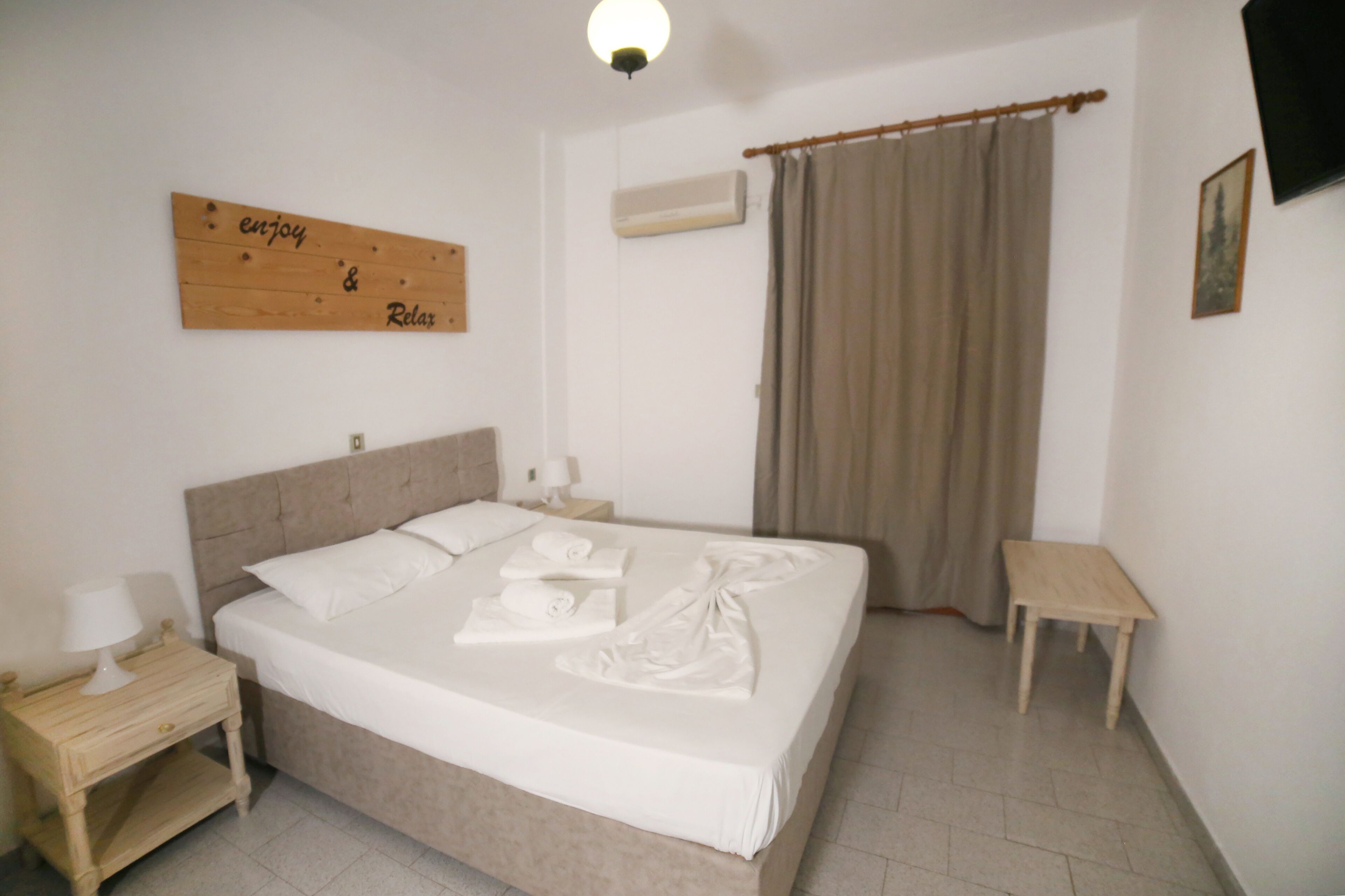 A neatly made bed with white linens and towels, a bedside table with a lamp, a wall-mounted air conditioner, and a wooden sign with the words "enjoy & relax".
