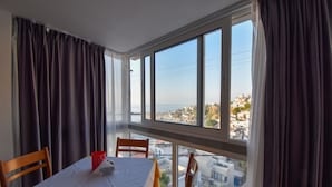 A room with a table set for two, large windows with curtains, and a view of a coastal town. - Kastro Hotel (Ikaria)