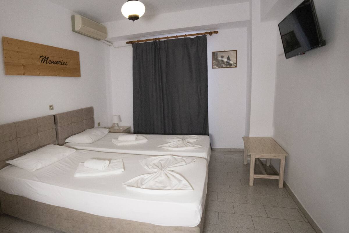 Habitación de hotel con dos camas, una mesita de madera, un televisor montado en la pared y un cartel que dice "Memories".