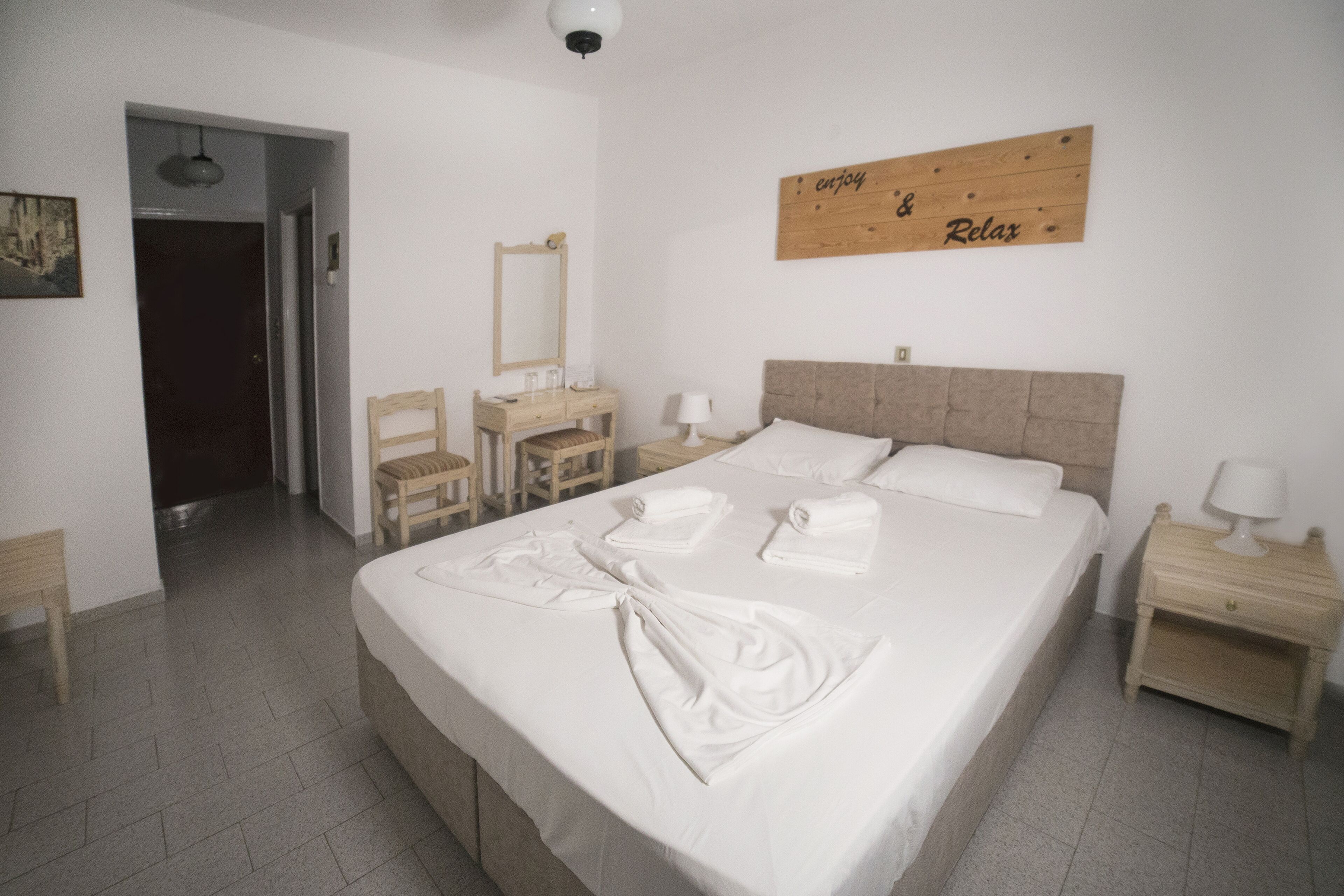 Una cama bien hecha con toallas, un cabecero de madera, una mesita de noche con lámpara y un letrero en la pared que dice "enjoy & relax".