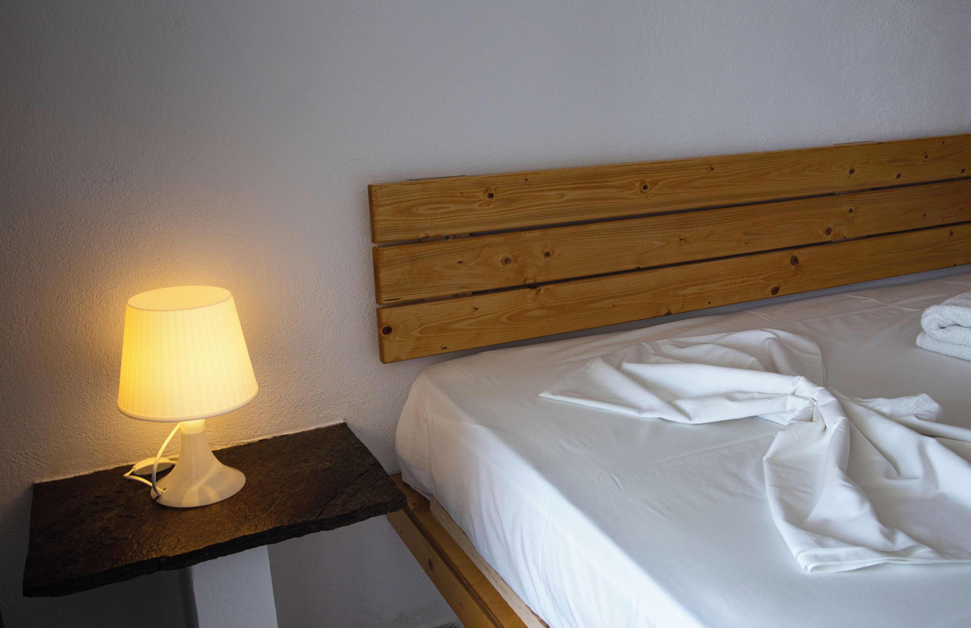 Een keurig opgemaakt bed met witte lakens en een houten hoofdbord. Een witte lamp staat op een donker houten nachtkastje naast het bed.