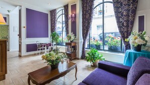 Une pièce avec un canapé violet, une table basse en bois et une fenêtre avec des rideaux. On y trouve des plantes en pot et une table ornée de fleurs.