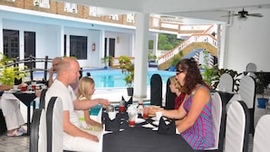 Uma área de refeições com mesas e cadeiras, uma piscina ao fundo e uma escadaria que leva a um andar superior.