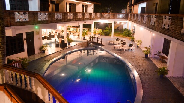 Área da piscina com espreguiçadeiras, uma pequena ponte e um espaço coberto com mesas e cadeiras.