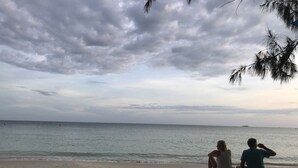 Two people sitting on a sandy beach with the ocean and a cloudy sky in the background. - Side Walk Boutique Hotel (Rayong)