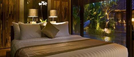 A bedroom with a large bed, two bedside lamps, and a view of a water feature outside.