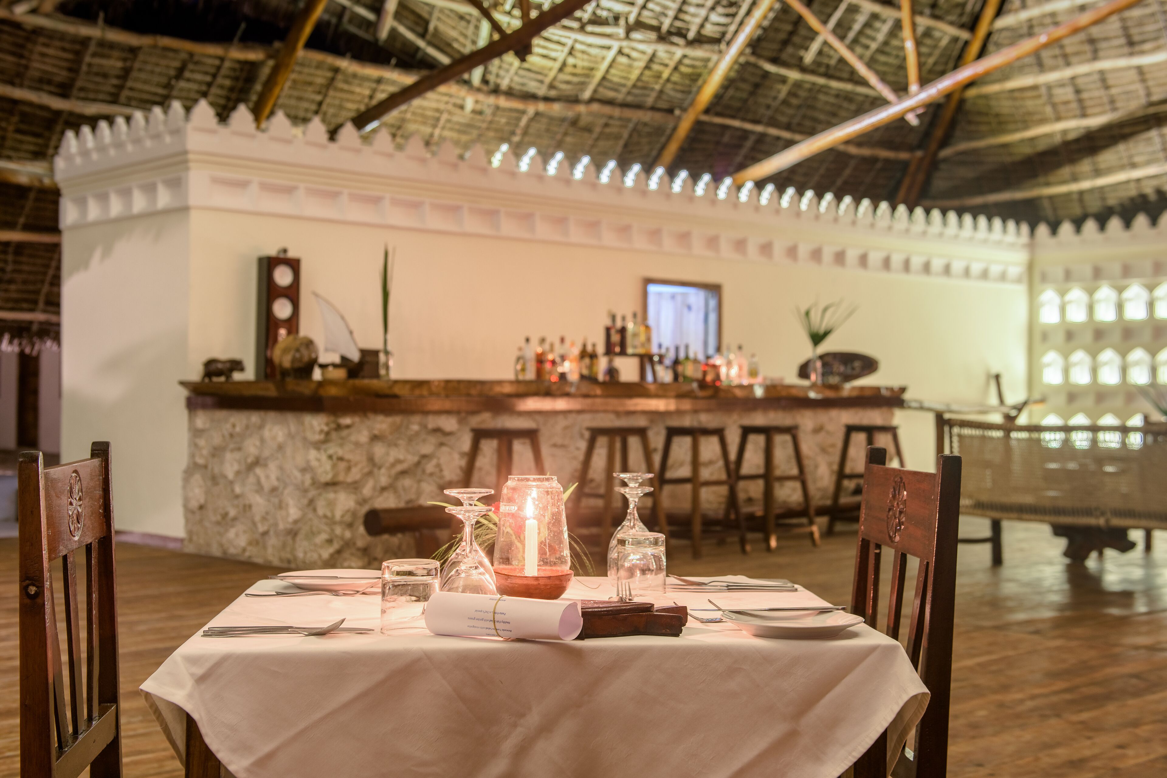 Un coin repas avec une table dressée pour deux personnes, un bar en arrière-plan et un plafond haut avec une toiture de chaume.