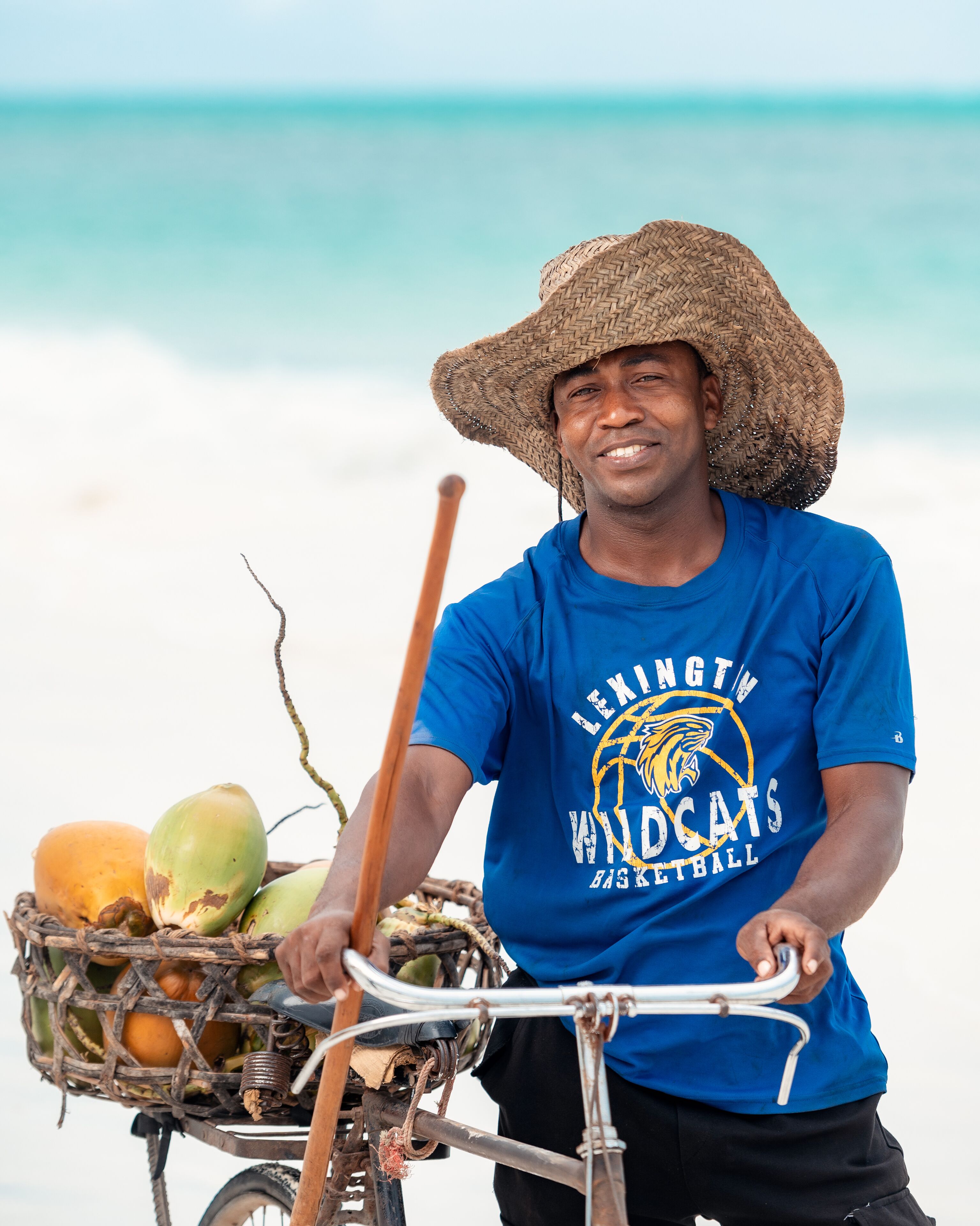 Uma pessoa de bicicleta, com um cesto de cocos e um bastão de madeira, vestindo uma camiseta azul com um logotipo e um chapéu de palha, em frente a uma praia.