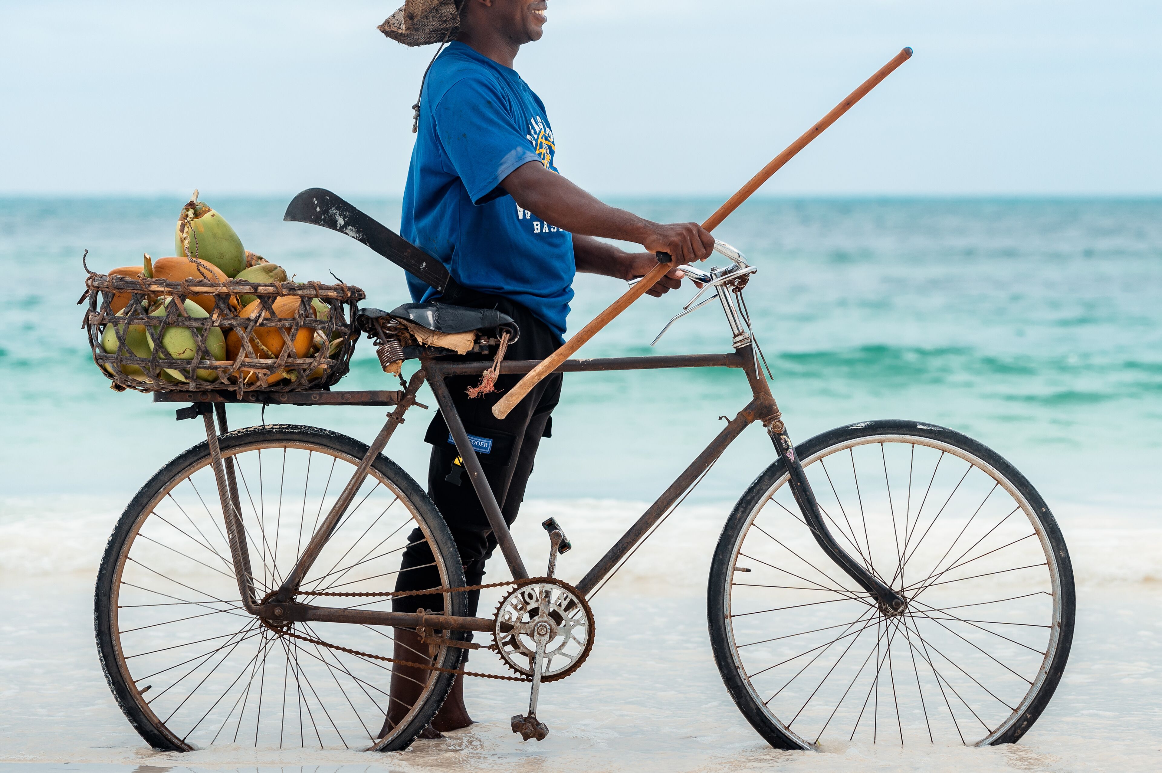 Un vélo avec un panier rempli de noix de coco, et une personne debout à côté, sur une plage.