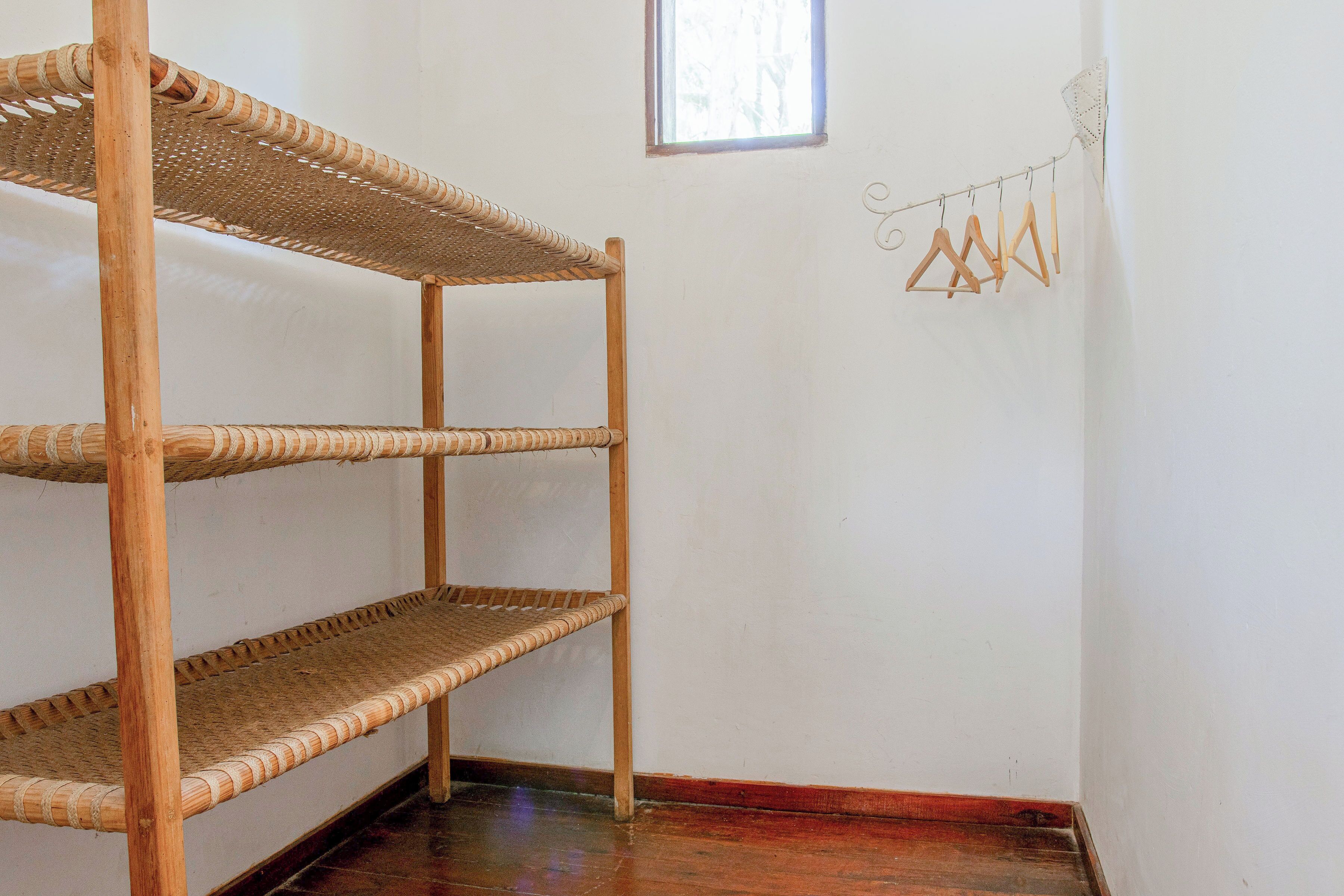 A corner of a room with a three-tiered wicker shelf unit against a white wall, a small window, and a wall-mounted hanger rod with wooden hangers.
