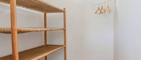 A corner of a room with a three-tiered wicker shelf unit against a white wall, a small window, and a wall-mounted hanger rod with wooden hangers.