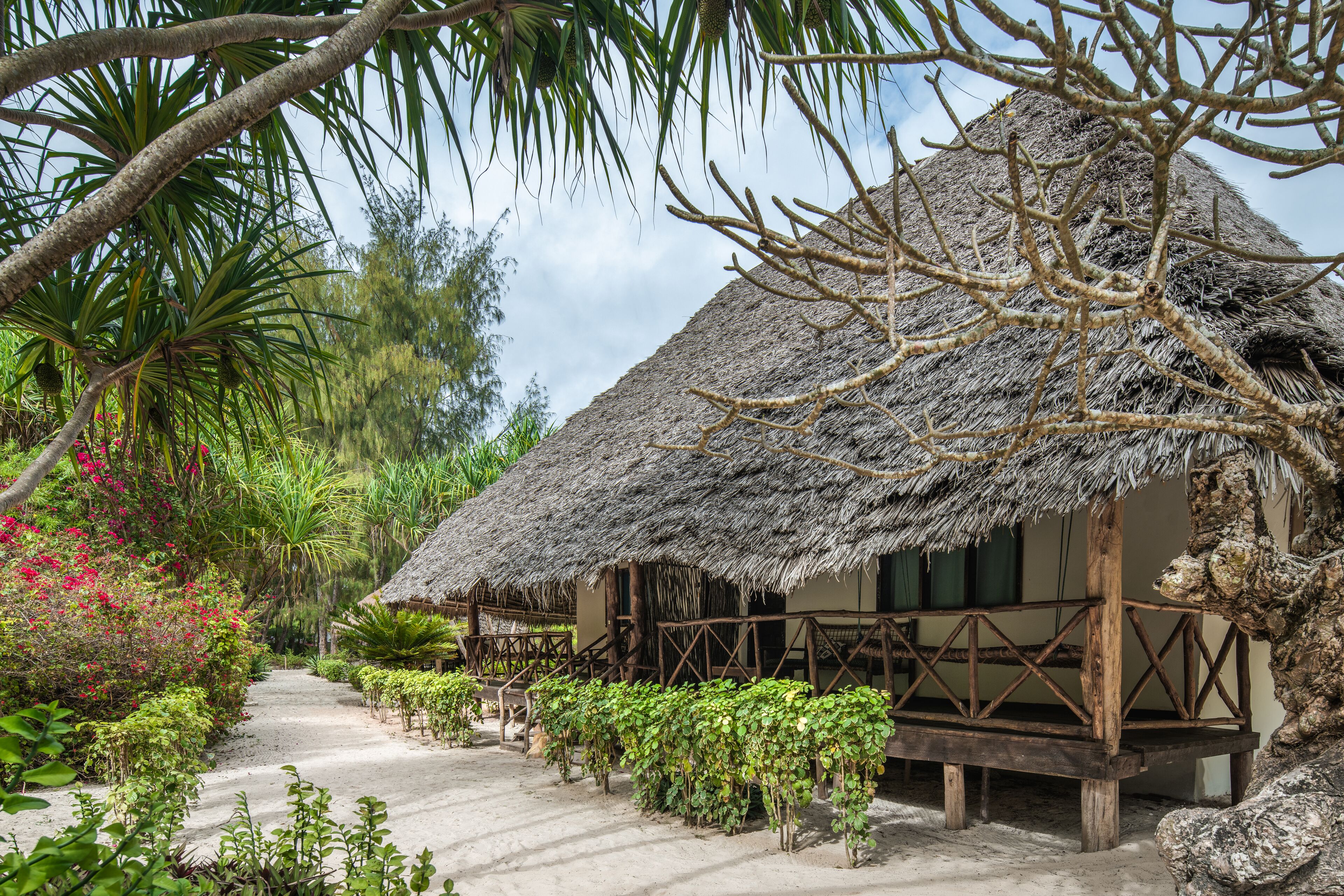 A thatched-roof house with a wooden porch, surrounded by lush greenery and a clear sky.