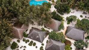 An aerial view of a resort with thatched-roof huts arranged in a grid pattern, surrounded by dense tropical vegetation and a swimming pool. - Hakuna Majiwe Beach Lodge (Paje)
